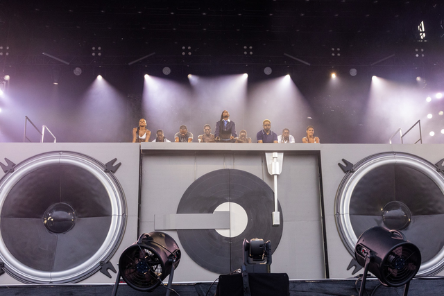 Un DJ y una tripulación se encuentran detrás de una configuración de gran escenario que se asemeja a un tocadiscos y altavoces, con brillantes fantelazos brillando arriba.